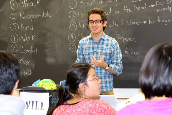 A young instructor stands confidently in front of a chalkboard, engaging a group of attentive teenagers.
