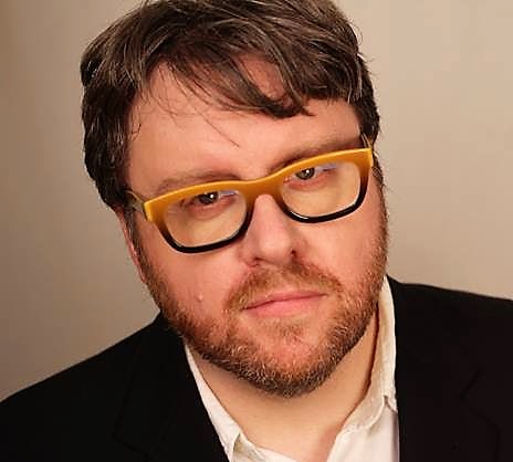 A bearded man wearing a black sports coat, white dress shirt and horn rimmed glasses. 