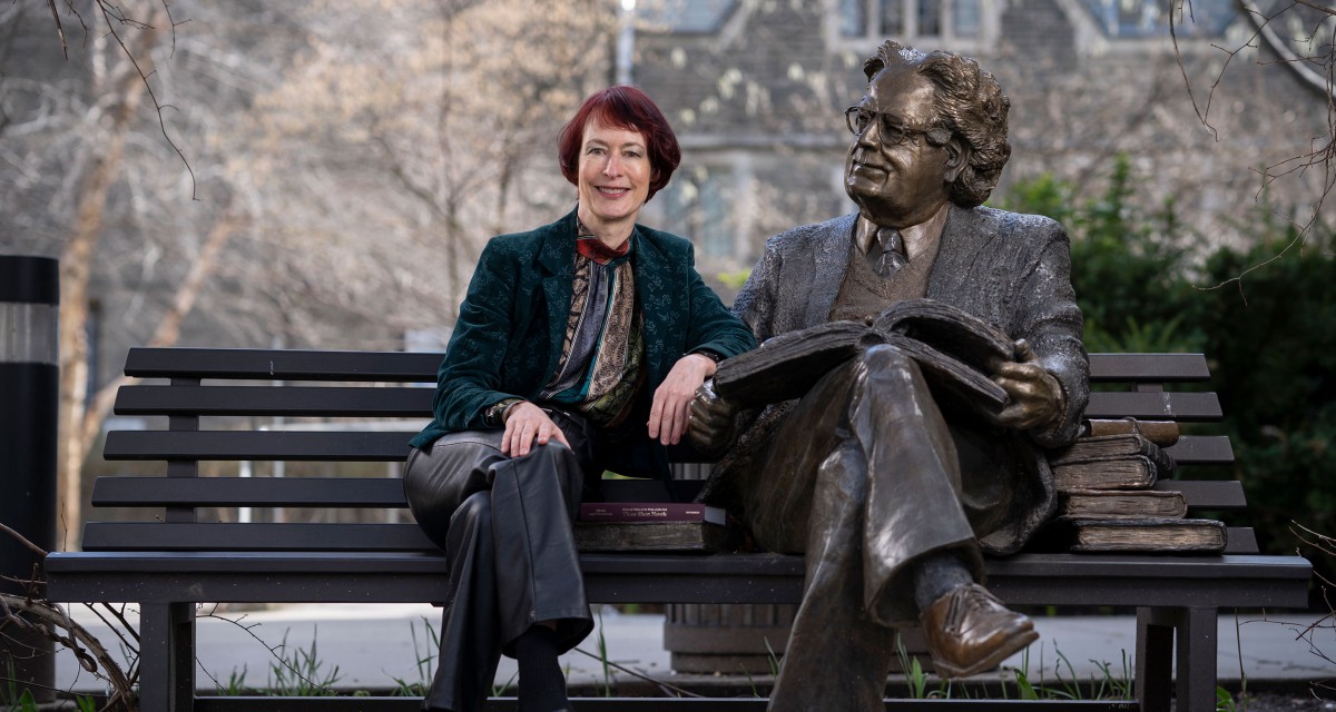 Principal Angela Esterhammer sits next to a bronze statue of Northrop Frye.