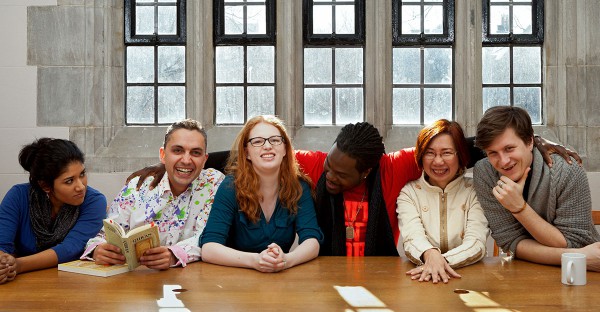 Six Emmanuel College students are seated around a rectangular wooden table, which is covered with books, papers, and laptops. They are all smiling and laughing, and their body language indicates that they are in good spirits. 